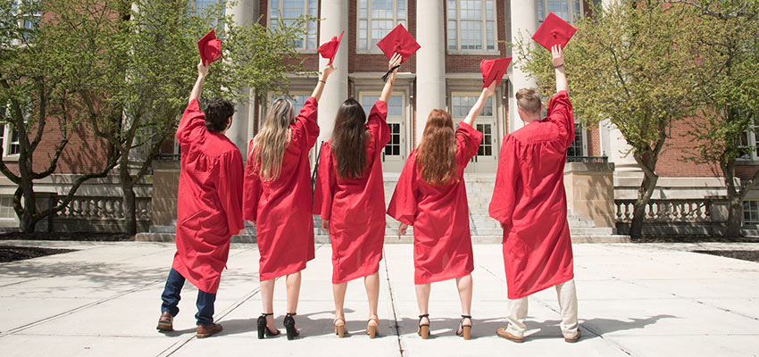Five graduates in red regalia stand facing Old Main Building, hold their caps high above their heads..