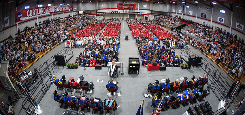Wide-angle view of the commencement ceremony, with faculty/staff in formal regalia and students in red, viewed from above center stage