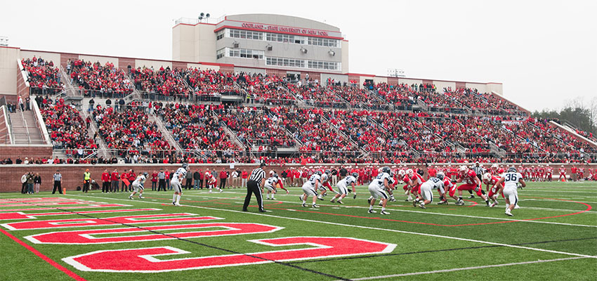 The Cortland Football team lines up against the Ithaca Bombers near the endzone at the line of scrimage, with bleachers in the background filled with fans dressed in red.
