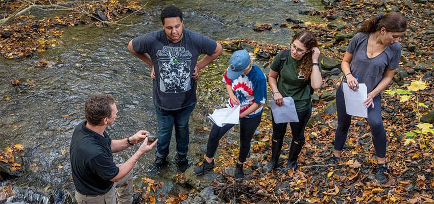 An instructor shows a rock to students at a creek bed as the students observe, holding paper and pens