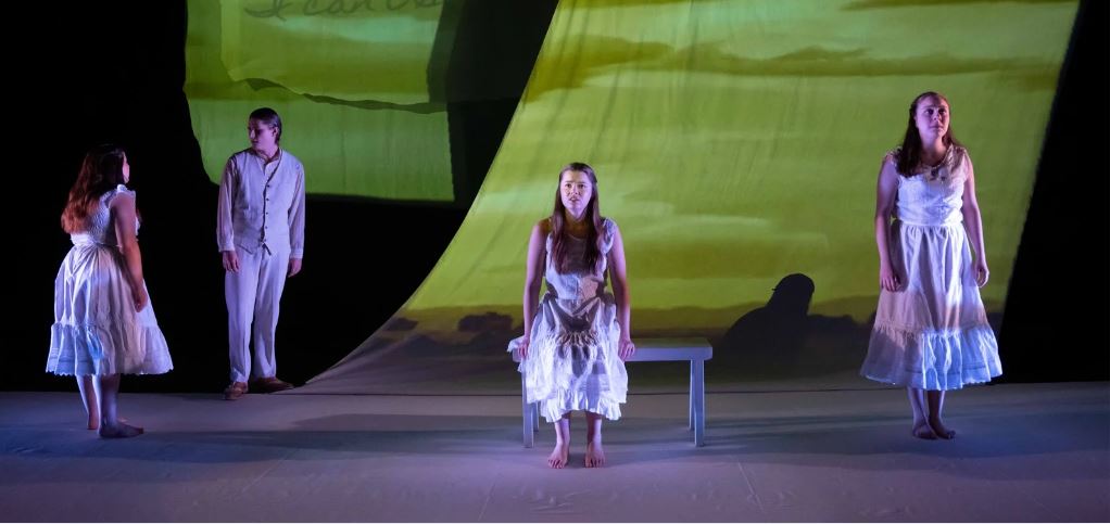 Women dresses in white stand spread across a dimly lit stage.