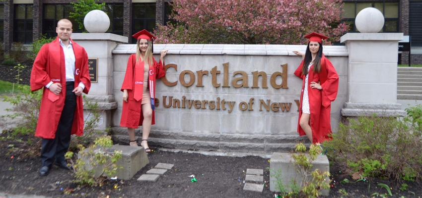 Students in graduation gowns standing in by the SUNY Cortland campus sign
