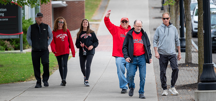 Family walking down campus, waving to camera with smiles.