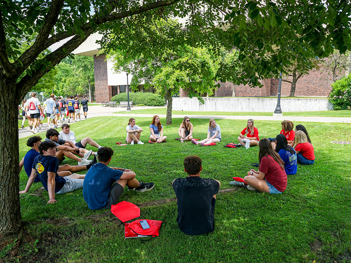 A group of students at Orientation sit in a circle on the grass outside Sperry Center
