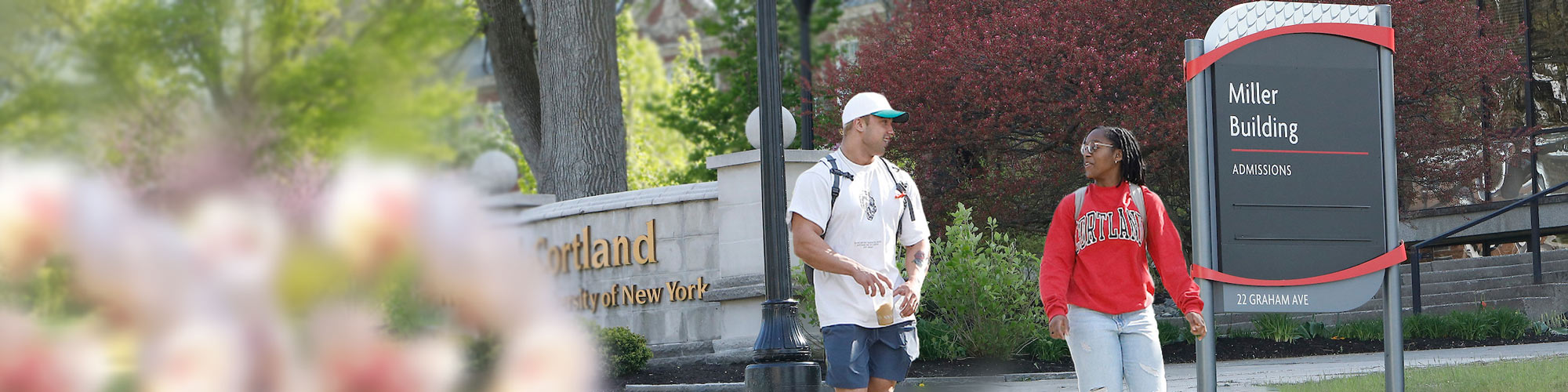 Two students walk together near the Cortland sign outside Miller Building on a spring day.