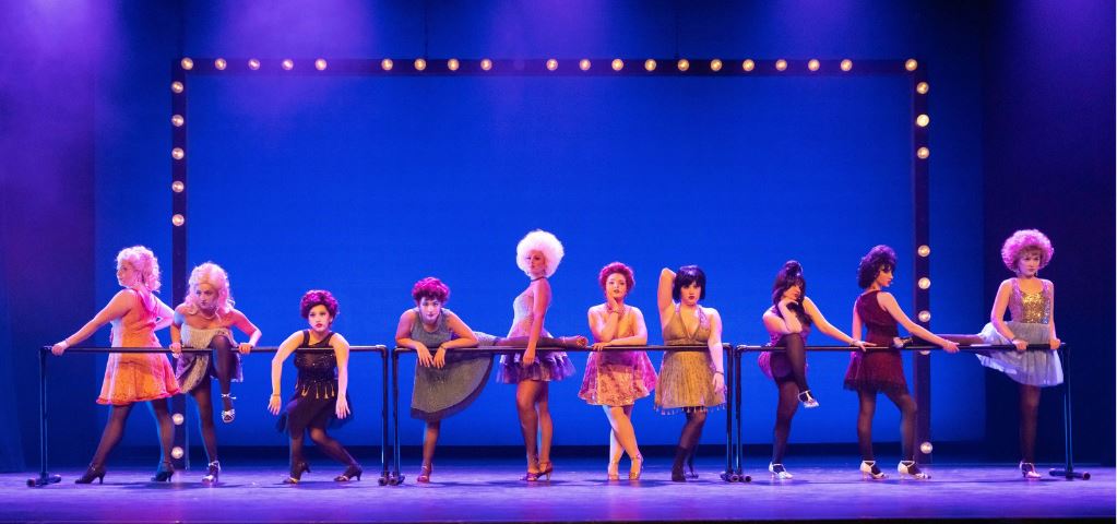 Ten women with big hair stand lines up behind a dance barre.