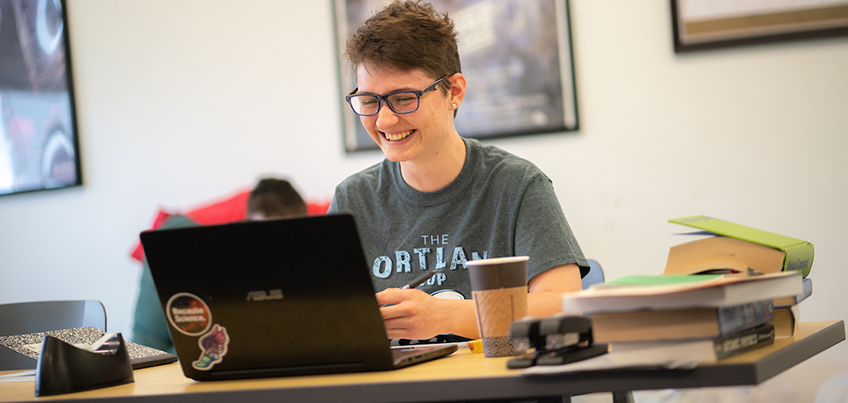 Student smiling and working on a laptop with a coffee nearby