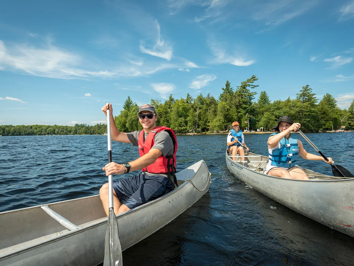 Students dressed in life jackets paddle in two canoes side by side on Raquette Lake with trees in the background