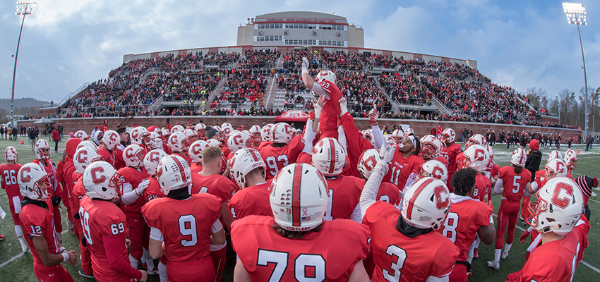 Cortland football player leaping in a huddle of teammates with the Cortland crowd in the background at the SUNY Cortland Stadium Complex
