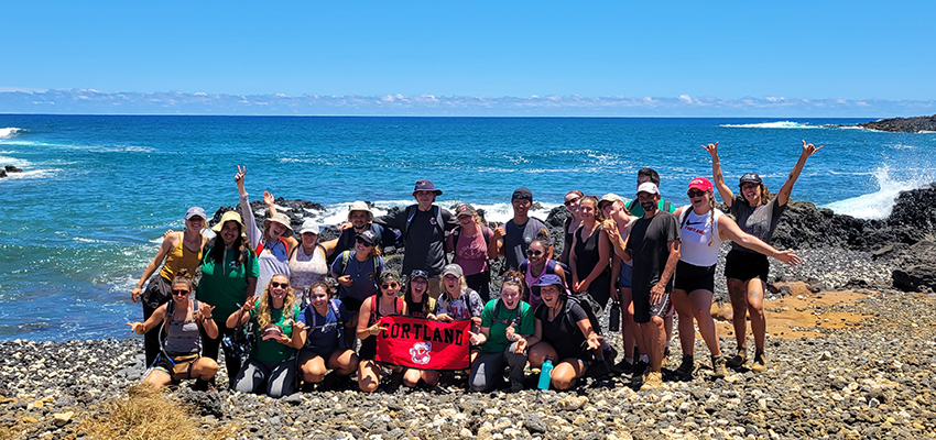 Students in ecotourism course pose near the ocean with a Cortland banner in Hawaii