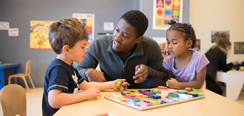 A male student teacher works with two youngsters to put together a puzzle