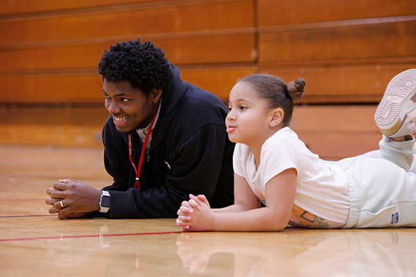 Makhatub Hassan smiles on the ground while teaching a physical education lesson with a young student seated nearby