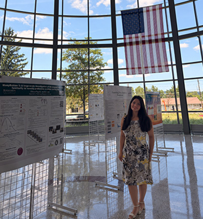 Student smiles and stands next to research poster with U.S. flag pictured in the background