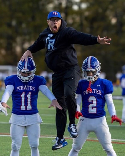 A youth football coach, Casey O'Keefe, jumps in the air between two youth football players posing in pads.
