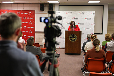 Sen. Lea Webb speaks at a podium with a SUNY Cortland backdrop