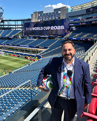 Greg Abdallah stands in a stadium with a scoreboard in the background and a soccer ball in his arm