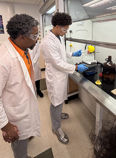 Photo of Dillian Vittini in a white lab coat, at right, conducting research in a chemistry lab with faculty member looking on