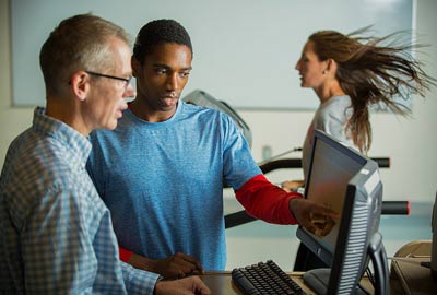 An instructor and student analyze data on a computer while a woman runs on a treadmill.