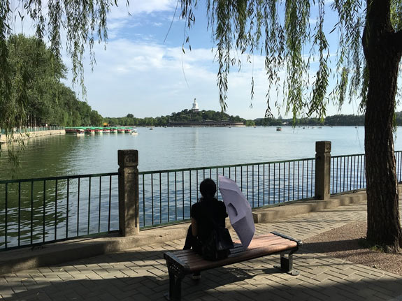 A person holds a sun umbrella while sitting on a bench by a park in Beijing, China